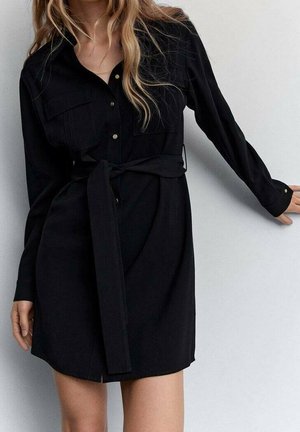 Femme portant une robe-chemise noire à manches longues avec des boutons, des poches poitrine et une ceinture en tissu nouée, debout contre un mur gris clair.