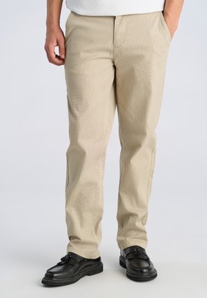 Uomo che indossa pantaloni beige a gamba dritta con mocassini neri con nappe, in piedi con una mano in tasca su sfondo semplice.