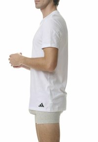 Biała bawełniana koszulka z krótkim rękawem, z czarnym logo Adidas umieszczonym w dolnym lewym rogu, widziana z boku.