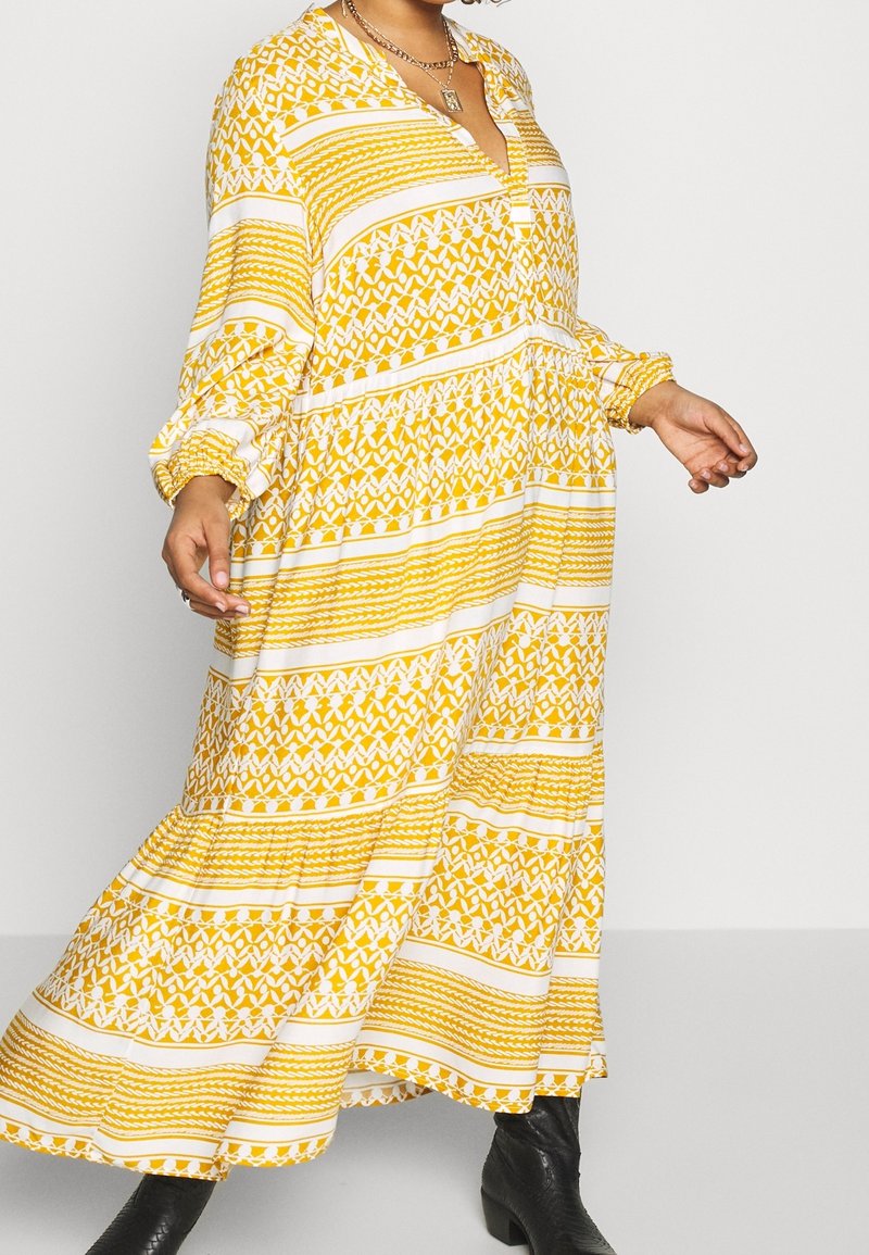 Kvinna klädd i en lång gul och vit mönstrad klänning med långa ärmar, svarta stövlar, ett halsband och en ring, stående mot en enkel bakgrund.