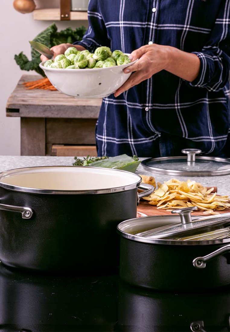 Schwarzer Kochtopf mit glänzendem Metalldeckel und weißem Innenraum; daneben ein kleinerer Topf mit Griff. Eine Schüssel mit Rosenkohl wird über einer Arbeitsplatte gehalten.