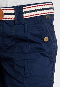 Pantalones azul marino con costuras detalladas, botones de latón con la etiqueta "edc" y un cinturón blanco con rayas rojas y azul marino.