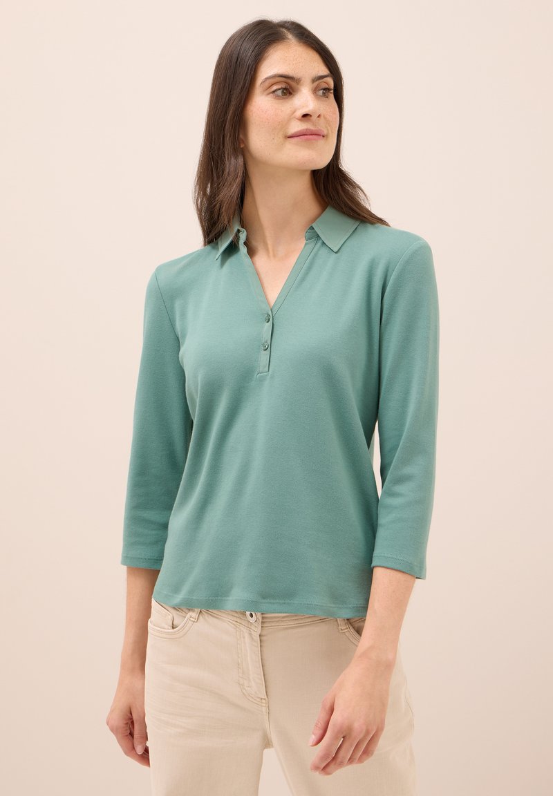 Lichtgroene polo met een kraag en drie knopen. Lange mouwen en een zachte textuur. Gecombineerd met lichtbeige broek.