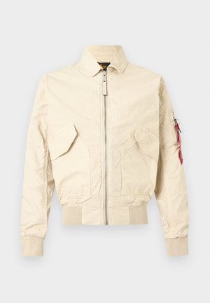 Creme-beige Bomberjacke mit frontalem Reißverschluss, zwei Klappentaschen, gerippten Bündchen und Saum sowie kleinem Reißverschlussfach am linken Ärmel mit rotem Etikett.