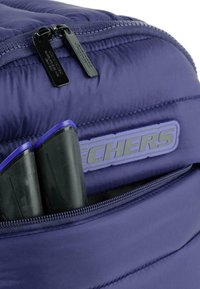 Mochila acolchada púrpura con dos cremalleras negras, un gran bolsillo frontal y un parche con el logo. El interior muestra artículos de almacenamiento negros con acentos en púrpura.