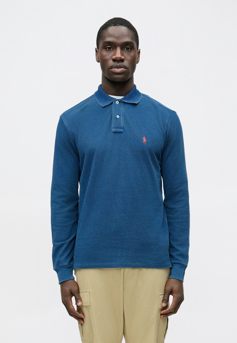 Modra polo majica z dolgimi rokavi in ovratnikom, z dvema gumboma in majhnim rdečim logotipom na levi strani prsnega koša. Mehka pletena tekstura.