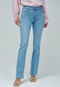 Jeans de mezclilla azul claro de corte acampanado con una cintura de altura media, diseño de cinco bolsillos y detalles de costura sutiles. Combinados con sandalias con tiras.