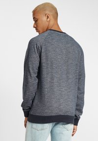 Tengerészkék pulóver texturált kötésmintával, bordás nyakkivágással és mandzsettákkal, valamint laza szabással. Látható varrás részletek a hátsó részen.