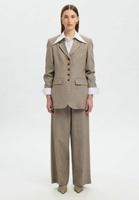 Completo beige su misura con pantaloni a gamba larga e blazer strutturato. Presenta un colletto prominente, cinque bottoni marroni e polsini a righe.