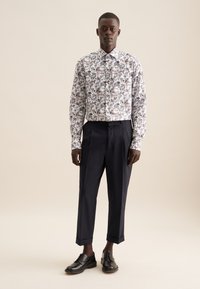 Witte kraagshirt met bloemenpatroon in verschillende tinten blauw, rood en groen. Gecombineerd met donkerblauwe op maat gemaakte broek en zwarte leren schoenen.
