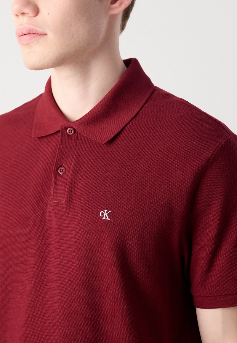 Burgundowa koszulka polo z teksturowanej tkaniny, z kołnierzykiem i zapięciem na dwa guziki, z małym białym logo na piersi.