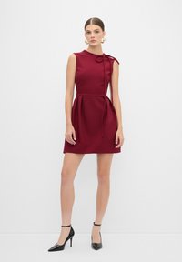 Abito senza maniche bordeaux con corpetto aderente, dettaglio alla spalla con fiocco e gonna svasata; abbinato a décolleté neri con punta a maggio.