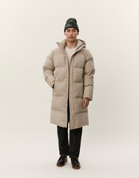 Uomo che indossa un lungo piumino beige, pantaloni neri, scarpe marrone scuro e un berretto verde, in piedi con una mano in tasca.