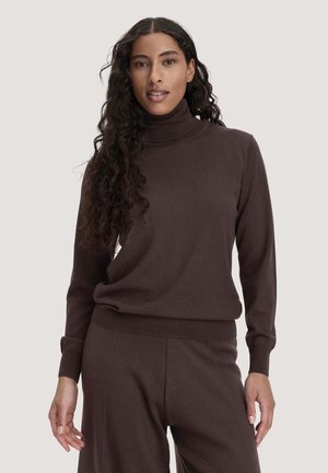 Vrouw met lang krullend haar die een donkerbruine coltrui en bijpassende broek draagt, staand tegen een effen lichte achtergrond.