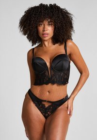 Vrouw met krullend haar in zwarte kanten lingerie, staande met één hand op haar heup tegen een eenvoudige grijze achtergrond.