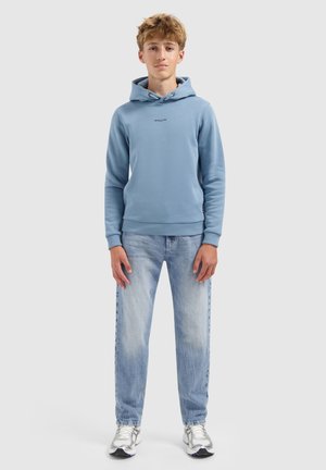 Tienerjongen staat, draagt een lichtblauwe hoodie met het logo "BALLIN", vervaagde blauwe spijkerbroek en witte sneakers, op een effen grijze achtergrond.