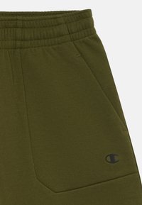 Pantalons de survêtement vert olive en mélange de coton, avec une taille élastique et une poche portant un logo noir sur le côté. Texture lisse.
