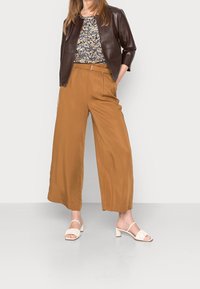 Giacca di pelle marrone, top a stampa floreale, pantaloni a gamba larga color sabbia e sandali con tacco beige. Texture lisce e vestibilità rilassata.