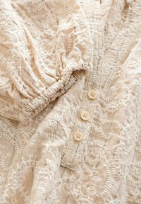 Chemise beige brodée avec des manches courtes bouffantes, ornée de motifs floraux blancs, comportant une patte de boutonnage avec trois boutons beiges et un tissu texturé.