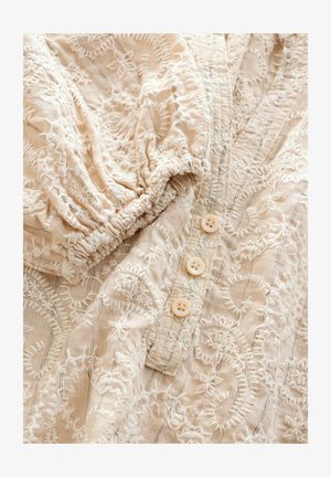 Camicia beige ricamata con maniche corte a sbuffo, dotata di motivi floreali bianchi, un'apertura con tre bottoni beige e tessuto testurizzato.