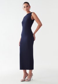 Jūras spēku zilas krāsas bez piedurknēm midi kleita ar pieguļošu siluetu, augstu apkakli un gludu tekstūru. Kombinēta ar metāliskiem papēžiem.
