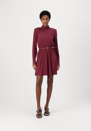 Robe bordeaux à manches longues avec un corsage froncé, un col haut et une ceinture assortie. Tissu lisse, longueur genoux, accompagnée de sandales noires.
