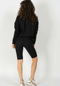 Czarny długi rękaw bluzka w połączeniu z czarnymi prążkowanymi szortami rowerowymi, prezentującymi gładką teksturę i luźny kształt. Białe i złote buty sportowe dopełniają stylizację.