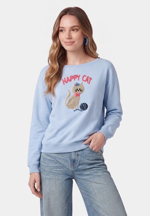 Giovane donna che indossa una felpa azzurro chiaro con un'illustrazione di gatto, le parole "Happy Cat," e una matassa di lana, abbinata a jeans blu.