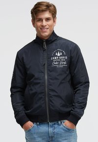 Marineblaue Bomberjacke mit gerippten Bündchen und Saum, ausgestattet mit einem durchgehenden Reißverschluss, grafischem Text und Segelmotive auf der Brust sowie Seitentaschen.