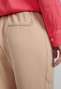 Beiga byxor i mjukt material med en sidoficka som har en guldknytt. Kombineras med en korallfärgad långärmad skjorta som visar en slät textur.