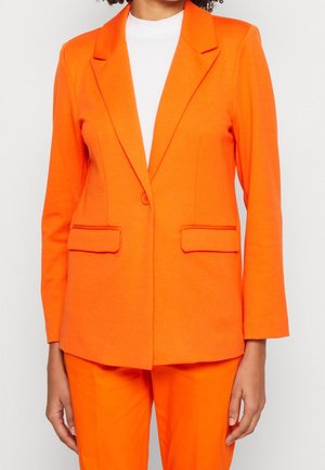 Blazer femme orange vif avec pantalon assorti, porté sur un haut blanc à col montant, présenté de la moitié du torse aux cuisses.