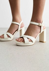 Sandalias blancas de tacón con tiras anchas entrelazadas y una hebilla en el tobillo. Cuentan con un tacón grueso y un acabado suave y texturizado.