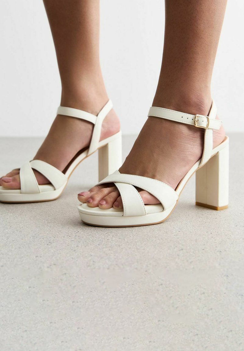 Sandalias blancas de tacón con tiras anchas entrelazadas y una hebilla en el tobillo. Cuentan con un tacón grueso y un acabado suave y texturizado.