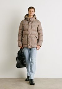 Casaco puffer listrado em marrom com capuz, apresentando um fecho de zíper e bolsos laterais, combinado com jeans claros e uma bolsa preta. Calçado preto.