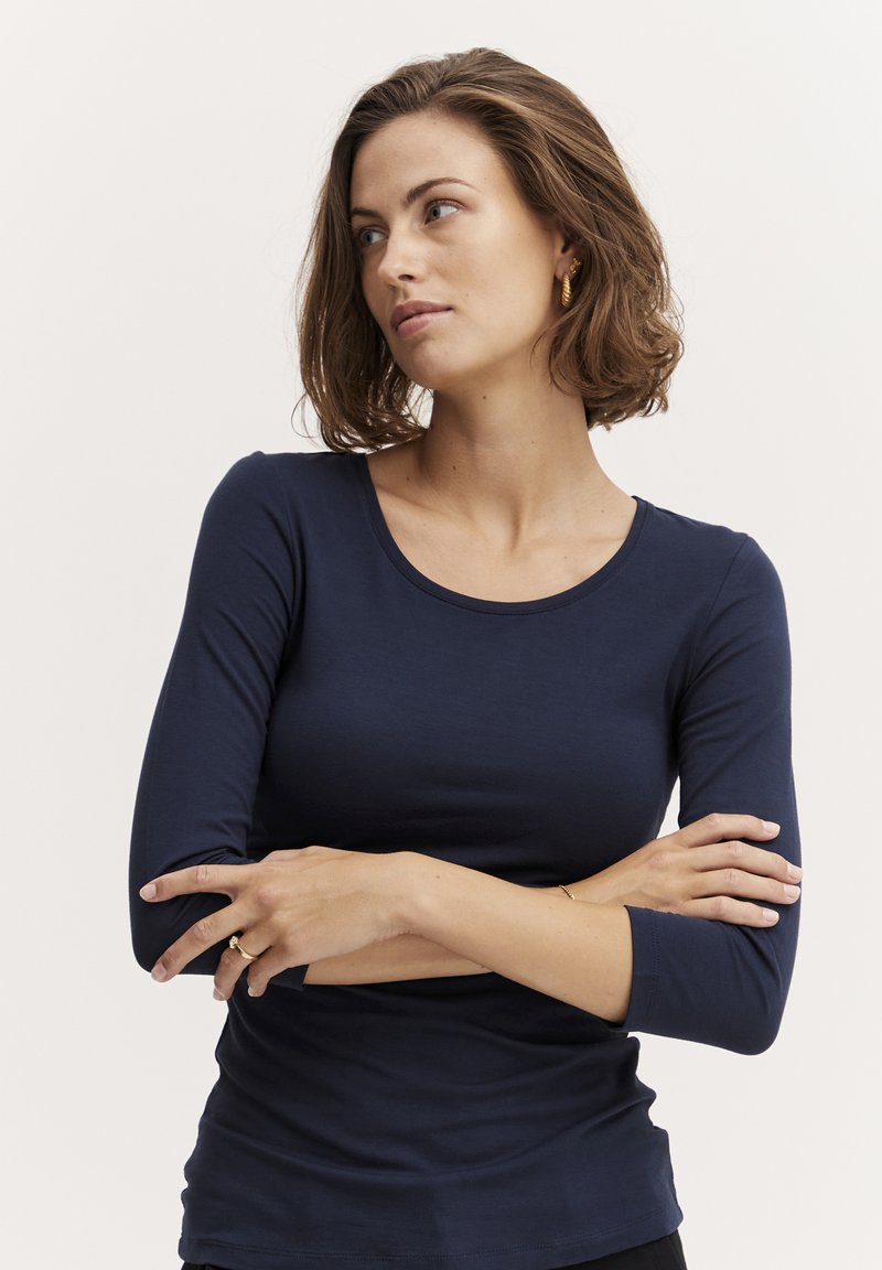 Bluemarin, bluză cu mânecă lungă, decolteu rotund, textură fină și croială ajustată. Modelul are brațele încrucișate și poartă bijuterii aurii discrete.