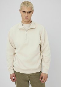 Sweatshirt beige clair à zip quart avec une texture douce, des poignets et un ourlet côtelés. Présente une fermeture éclair en métal et un design décontracté.