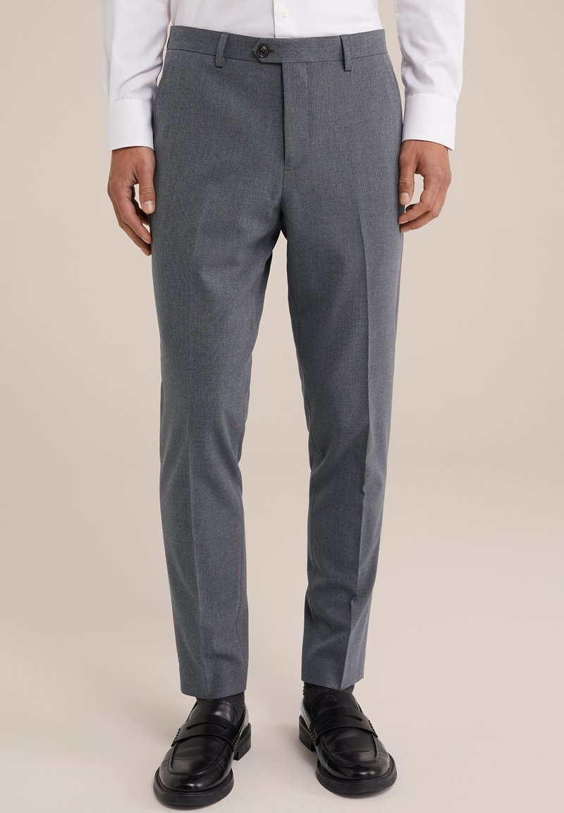 Homme portant un pantalon de costume gris slim-fit associé à des mocassins en cuir noirs et une chemise blanche à manches longues, debout devant un fond beige.