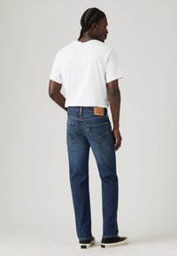 Jeans in denim blu con un taglio dritto, dotati di una patch posteriore in pelle e cuciture a contrasto. Indossati con una t-shirt bianca e scarpe nere.