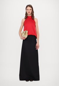 Haut sans manches rouge avec un col montant, associé à une longue jupe noire. Tient un sac à main doré texturé avec un détail en nœud.
