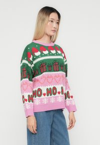 Ung kvinde iført en grøn og pink julesweater med julemandshætter, gaver, slikstænger, hjerter, teksten "HO HO HO" og snefnug.