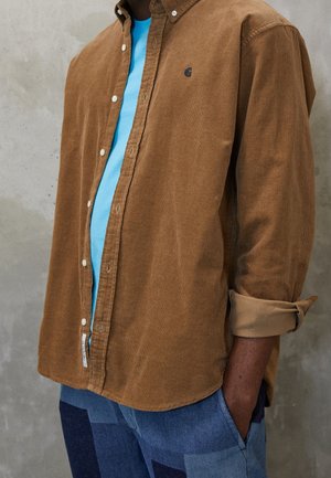 Person wearing a brown corduroy shirt over a light blue t-shirt and patchwork blue jeans, with one hand in pocket.