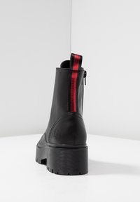 Botín de cuero negro hasta el tobillo con puntera redondeada, suela estriada y un bucle elástico con rayas rojas en la parte posterior para facilitar el uso. Detalle de cremallera lateral.