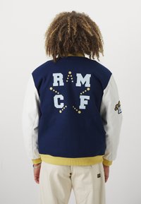 Mornarska in bela bomber jakna z velikimi svetlo modrimi črkami "RM" in "CF" na hrbtu, rumenimi detajli in rebrastim robom.
