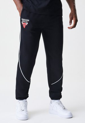 Black sweatpants featuring a red Chicago Bulls logo, white piping accents, and a tapered fit. Worn with white sneakers.