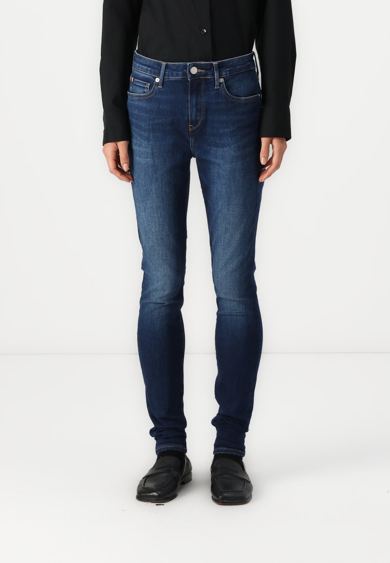 Person som har på seg mørkeblå skinny jeans, svart langermet topp og svarte loafers, står mot en hvit bakgrunn.