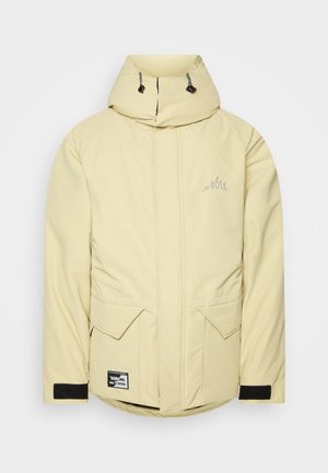 Homme portant une veste d’hiver beige à capuche sur un col roulé noir, un pantalon noir et des baskets blanches, debout devant un fond uni.