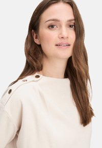 Jasnop beżowy sweter o prążkowanej fakturze, okrągłym dekolcie i ozdobnych guzikach na ramieniu. Miękka tkanina i luźny krój.