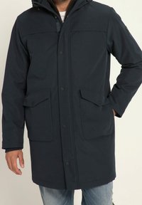 Imperméable marine avec capuche, boutons pression, deux poches avant et tissu légèrement texturé. Coupe décontractée avec une longueur mi-longue.
