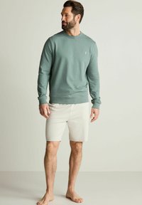 Sudadera verde clara con cuello redondo y puños de canalé, combinada con pantalones cortos beige. Corte relajado, tejido suave, sin patrones o logotipos visibles.