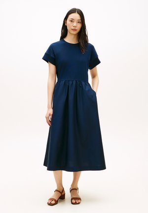 Vrouw staat in een marineblauwe midi-jurk met korte mouwen en een geplooide taille, gecombineerd met bruine sandalen met hak, tegen een neutrale achtergrond.